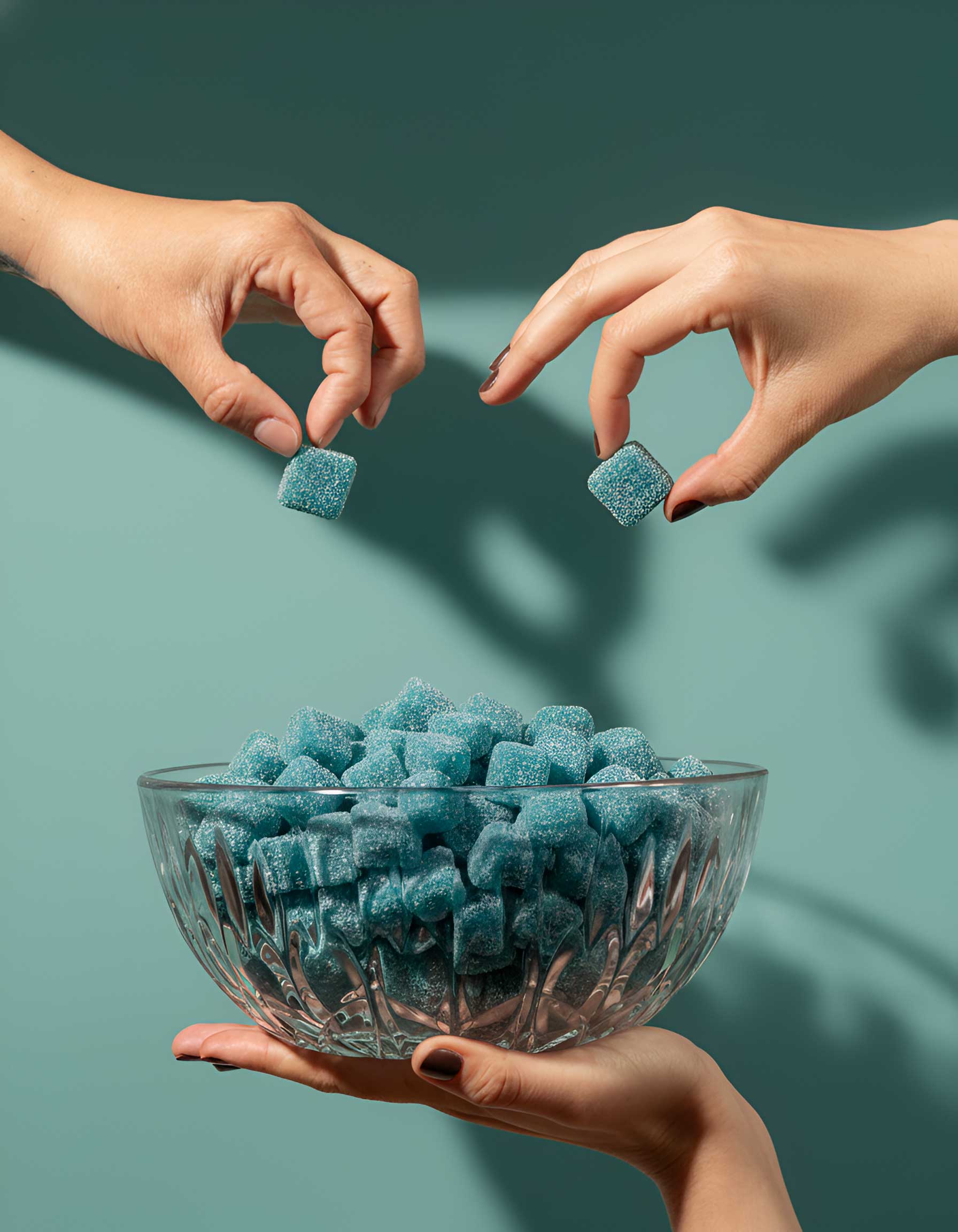 Three hands reaching towards a bowl of blue gummy cubes against a teal background