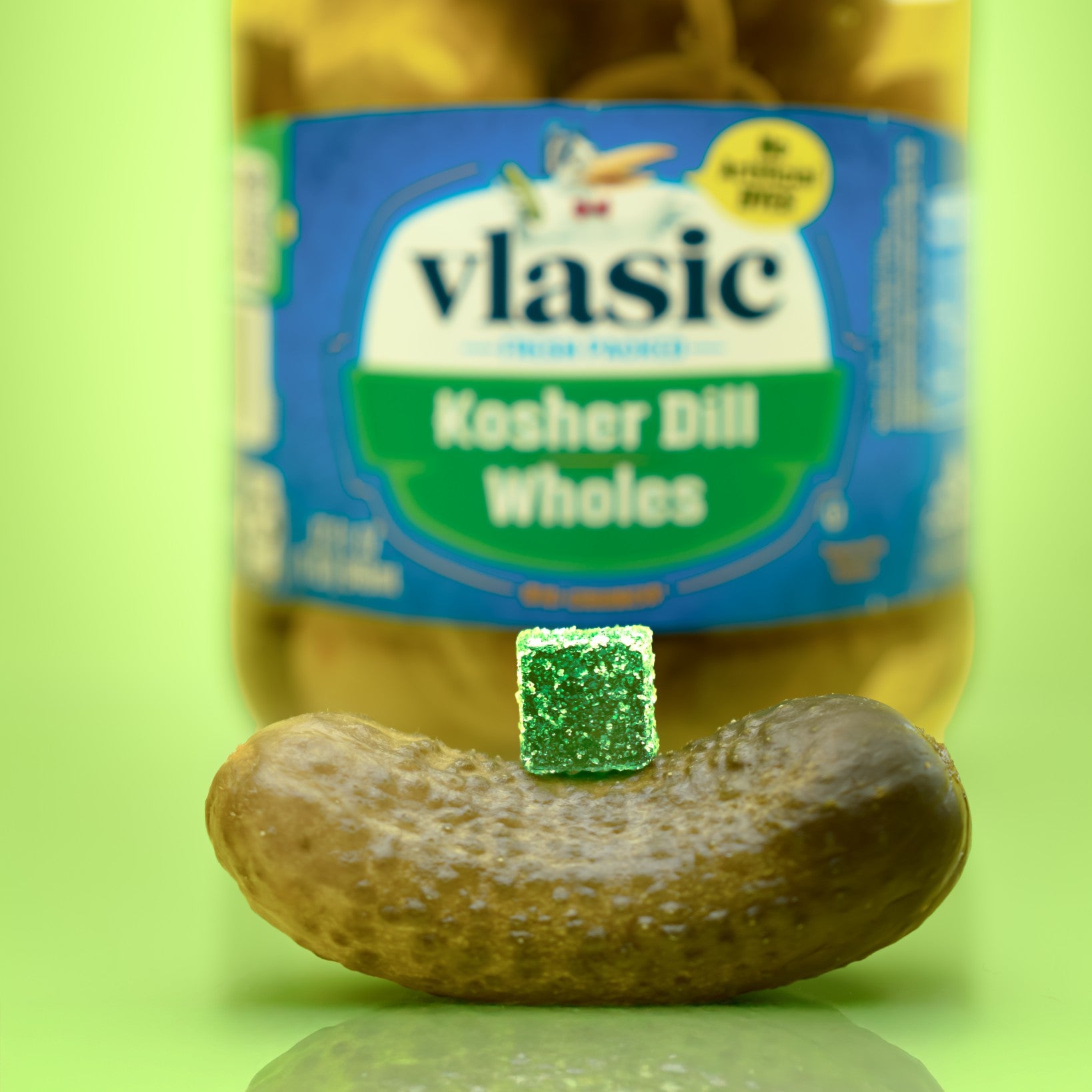 Jar of pickles with a pickle and pickle gummy in front on a green background